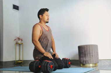 Evde dambıllarla egzersiz yaparken mola veren bir adam.