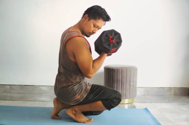 Kol halterleri kullanarak içeride egzersiz yapan motive olmuş bir birey. Bir ev ortamında kendini adamanın, sağlığın ve kendini geliştirmenin özünü yakalar.