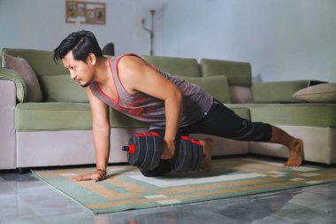 Adam evde bir dambılla güç alıştırması yapıyor. Kapalı bir spor salonu, rahat bir ev düzeni, evin rahatlığından sağlıklı ve aktif bir yaşam tarzına olan bağlılığını gösteriyor.