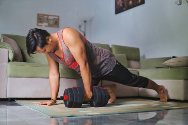 Adam evde bir dambılla güç alıştırması yapıyor. Kapalı bir spor salonu, rahat bir ev düzeni, evin rahatlığından sağlıklı ve aktif bir yaşam tarzına olan bağlılığını gösteriyor.