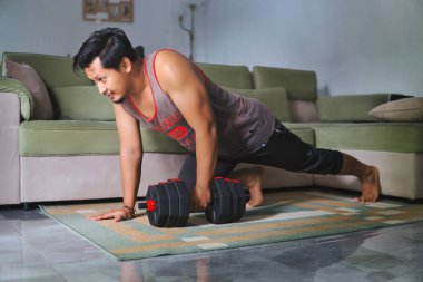 Adam evde bir dambılla güç alıştırması yapıyor. Kapalı bir spor salonu, rahat bir ev düzeni, evin rahatlığından sağlıklı ve aktif bir yaşam tarzına olan bağlılığını gösteriyor.