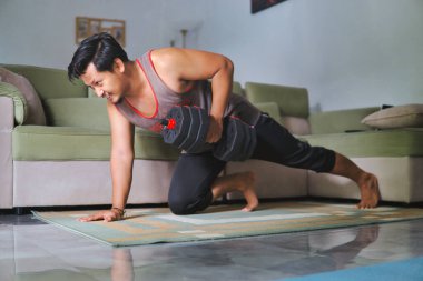 Adam evde bir dambılla güç alıştırması yapıyor. Kapalı bir spor salonu, rahat bir ev düzeni, evin rahatlığından sağlıklı ve aktif bir yaşam tarzına olan bağlılığını gösteriyor.
