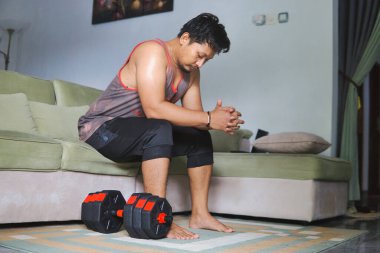 Oturma odasında halterle egzersiz yapan bir adam kanepeye oturur ve düşünür.