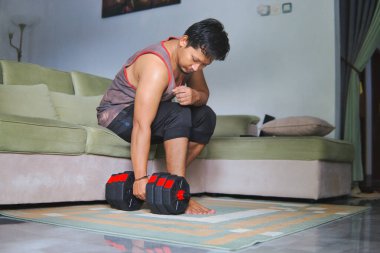 Oturma odasında bir dambılla egzersiz yapan bir adam, rahat ve rahat bir ev ortamında spor ve sağlığa olan bağlılığını yansıtır.