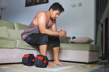 Oturma odasında halterle egzersiz yapan bir adam kanepeye oturur ve düşünür.