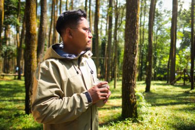 Adam sakin bir orman ortamında elinde sıcak bir içecekle, doğal ortamda duruyor ve çevredeki sükunetin tadını çıkarıyor, kişisel rahatlık ve farkındalığın altını çiziyor.