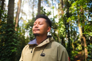 Bir adamın yemyeşil bir ormanda sükuneti kucakladığı sakin bir an, güneş ışığı tepe örtüsünün içinden süzülüyor, doğanın güzelliğini ve dışarıdaki sessizliği vurguluyor.