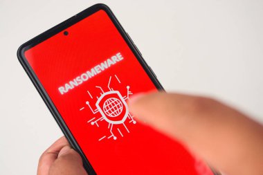 Ekranda gösterilen fidye virüsü uyarısı ile akıllı telefonu tutan bir elin yakın çekim görüntüsü siber güvenlik ve kötü niyetli yazılımın etrafındaki endişeleri vurguluyor..
