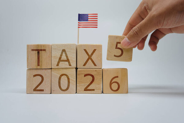 Close-up of a hand changing wooden blocks to display TAX 2026 with a miniature USA flag on top. Concept for United States taxation, fiscal policy, and financial planning for 2026.
