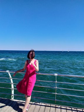 Açık pembe bir yaz elbisesi içindeki genç bir kadın, saçları rüzgarda uçuşan bir iskelede duruyor. Derin mavi okyanus ufka doğru uzanıyor. Güneşli hava, canlı renkler ve rahat duruş kaygısız bir yaz havası veriyor..
