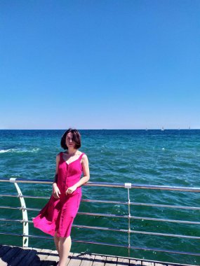 Açık pembe bir yaz elbisesi içindeki genç bir kadın, saçları rüzgarda uçuşan bir iskelede duruyor. Derin mavi okyanus ufka doğru uzanıyor. Güneşli hava, canlı renkler ve rahat duruş kaygısız bir yaz havası veriyor..