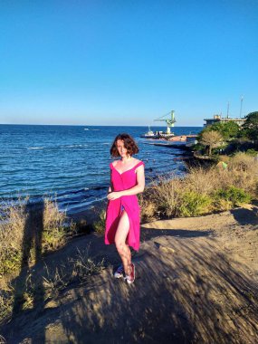 Açık pembe keten elbise giyen genç bir kadın deniz kenarındaki ahşap iskelede kameraya bakıyor. Rüzgar nazikçe elbisesini ve saçlarını hareket ettiriyor ve engin mavi gökyüzü ve okyanus sakin ve açık bir atmosfer yaratıyor. Ufukta yelkenliler görünüyor.