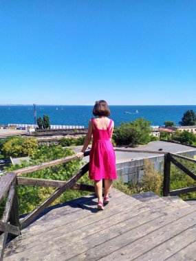 Açık pembe keten elbise giyen genç bir kadın deniz kenarındaki ahşap iskelede kameraya bakıyor. Rüzgar nazikçe elbisesini ve saçlarını hareket ettiriyor ve engin mavi gökyüzü ve okyanus sakin ve açık bir atmosfer yaratıyor. Ufukta yelkenliler görünüyor.