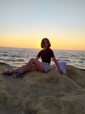 Gün doğumunda deniz kenarında kayaların üzerinde oturan, elinde havadaki bir kumaşı tutan bir kadının silueti. Gün doğumunda deniz kenarında elinde bir kumaşla oturan bir kadının görüntüsü. 