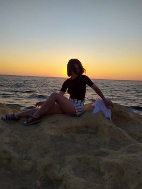 Gün doğumunda deniz kenarında kayaların üzerinde oturan, elinde havadaki bir kumaşı tutan bir kadının silueti. Gün doğumunda deniz kenarında elinde bir kumaşla oturan bir kadının görüntüsü. 