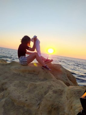 Gün doğumunda deniz kenarında kayaların üzerinde oturan, elinde havadaki bir kumaşı tutan bir kadının silueti. Gün doğumunda deniz kenarında elinde bir kumaşla oturan bir kadının görüntüsü. 