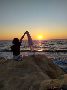 Gün doğumunda deniz kenarında kayaların üzerinde oturan, elinde havadaki bir kumaşı tutan bir kadının silueti. Gün doğumunda deniz kenarında elinde bir kumaşla oturan bir kadının görüntüsü.