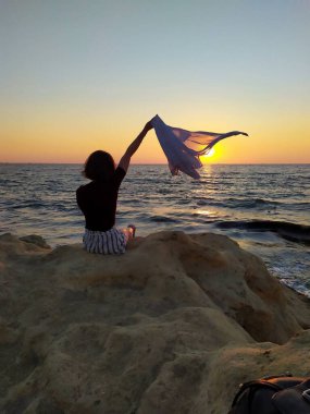 Gün doğumunda deniz kenarında kayaların üzerinde oturan, elinde havadaki bir kumaşı tutan bir kadının silueti. Gün doğumunda deniz kenarında elinde bir kumaşla oturan bir kadının görüntüsü.