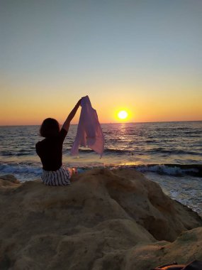 Gün doğumunda deniz kenarında kayaların üzerinde oturan, elinde havadaki bir kumaşı tutan bir kadının silueti. Gün doğumunda deniz kenarında elinde bir kumaşla oturan bir kadının görüntüsü. 