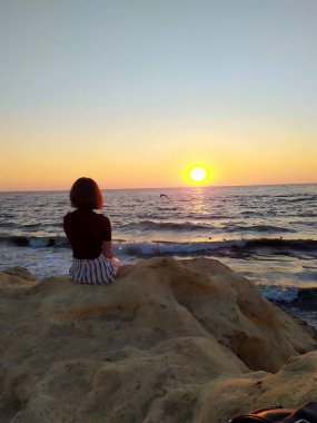 Gün doğumunda deniz kenarında kayaların üzerinde oturan, elinde havadaki bir kumaşı tutan bir kadının silueti. Gün doğumunda deniz kenarında elinde bir kumaşla oturan bir kadının görüntüsü. 