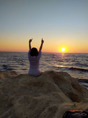 Gün doğumunda deniz kenarında kayaların üzerinde oturan, elinde havadaki bir kumaşı tutan bir kadının silueti. Gün doğumunda deniz kenarında elinde bir kumaşla oturan bir kadının görüntüsü.