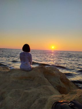 Gün doğumunda deniz kenarında kayaların üzerinde oturan, elinde bir kumaş tutan bir kadının silueti. Sahilde huzurlu bir gün doğumunun keyfini çıkaran turist kadın, uçurumlarda oturuyor ve okyanus manzarasına hayran. Seyahat, özgürlük ve rahatlama kavramı