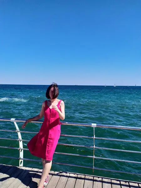 Açık pembe bir yaz elbisesi içindeki genç bir kadın, saçları rüzgarda uçuşan bir iskelede duruyor. Derin mavi okyanus ufka doğru uzanıyor. Güneşli hava, canlı renkler ve rahat duruş kaygısız bir yaz havası veriyor..