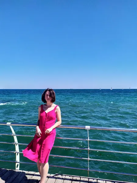 Açık pembe bir yaz elbisesi içindeki genç bir kadın, saçları rüzgarda uçuşan bir iskelede duruyor. Derin mavi okyanus ufka doğru uzanıyor. Güneşli hava, canlı renkler ve rahat duruş kaygısız bir yaz havası veriyor..