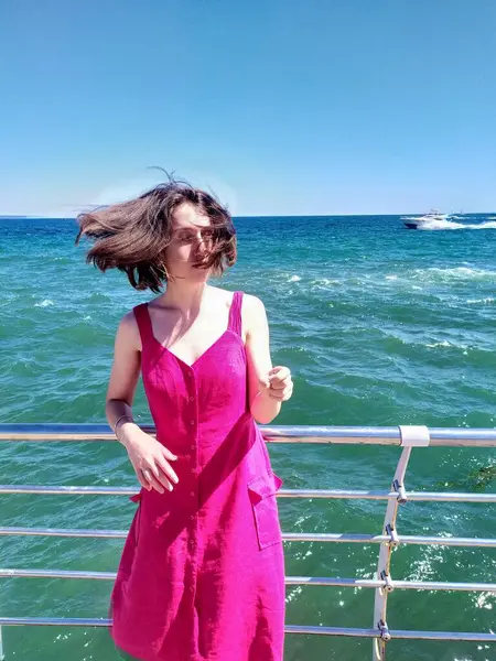 Açık pembe keten elbise giyen genç bir kadın deniz kenarındaki ahşap iskelede kameraya bakıyor. Rüzgar nazikçe elbisesini ve saçlarını hareket ettiriyor ve engin mavi gökyüzü ve okyanus sakin ve açık bir atmosfer yaratıyor. Ufukta yelkenliler görünüyor.