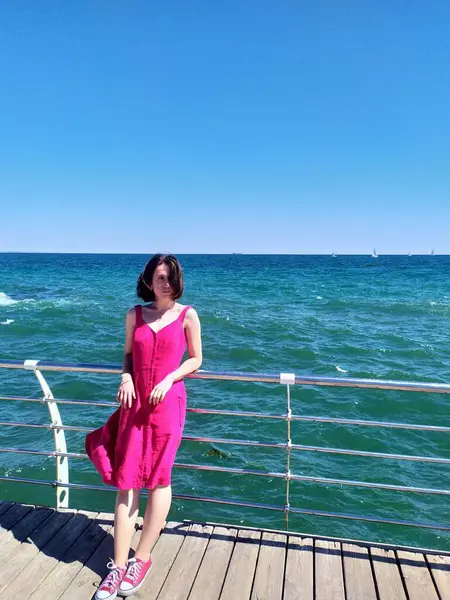 Açık pembe keten elbise giyen genç bir kadın deniz kenarındaki ahşap iskelede kameraya bakıyor. Rüzgar nazikçe elbisesini ve saçlarını hareket ettiriyor ve engin mavi gökyüzü ve okyanus sakin ve açık bir atmosfer yaratıyor. Ufukta yelkenliler görünüyor.