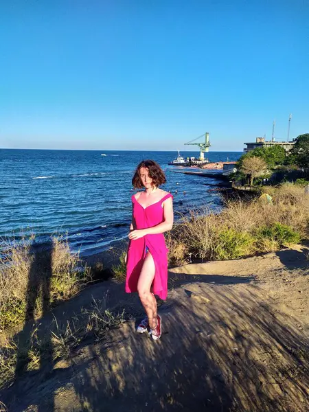 Açık pembe keten elbise giyen genç bir kadın deniz kenarındaki ahşap iskelede kameraya bakıyor. Rüzgar nazikçe elbisesini ve saçlarını hareket ettiriyor ve engin mavi gökyüzü ve okyanus sakin ve açık bir atmosfer yaratıyor. Ufukta yelkenliler görünüyor.