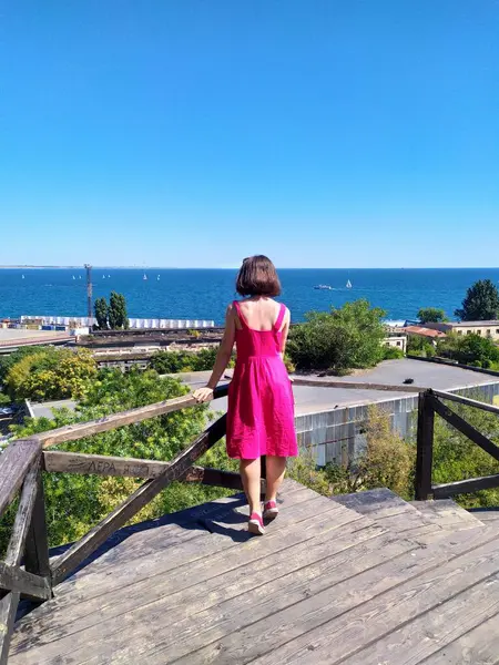 Açık pembe keten elbise giyen genç bir kadın deniz kenarındaki ahşap iskelede kameraya bakıyor. Rüzgar nazikçe elbisesini ve saçlarını hareket ettiriyor ve engin mavi gökyüzü ve okyanus sakin ve açık bir atmosfer yaratıyor. Ufukta yelkenliler görünüyor.