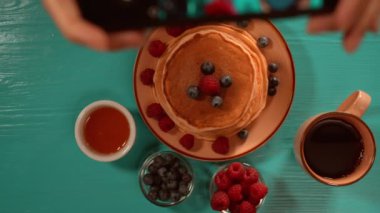 Kırmızı böğürtlen ve bal olan sıcak kekler mavi bir masanın üzerinde, bir kişi akıllı telefonuyla çanağın fotoğraflarını çekerken,.