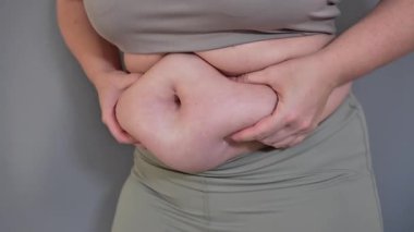 Karnını sıkan, doğal vücut kıvrımlarını ortaya çıkaran bir kadının yakın çekimi. Gri arka planda spor giyer. Görüntü vücudun pozitifliğini ve kendini kabulünü vurguluyor.
