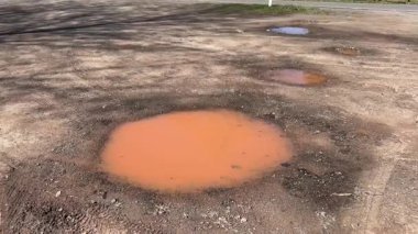 Parlak gökyüzünün altında, pürüzsüz toprak yol yüzeyine dağılmış, yumuşak gölgeler ve kırsal atmosfer oluşturan ağaçlarla çevrili, çok büyük, çamurlu su birikintileri.