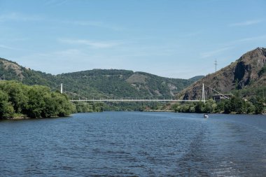 Yeşil tepeler ve parlak mavi gökyüzü ile çerçevelenmiş, doğal güzellik ve sükuneti gösteren bir nehir manzarası.