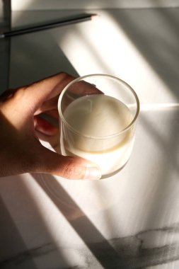 Bir bardak süt dokunuşu ve arkasında koyu gölgeli bulanık bir defter ve kalem. Öğleden sonra atıştırması, sağlıklı yiyecekler, içecek, doğal ışık, güneş ışığı, ev, arka plan için sıcak hava konsepti. Yaz boyunca natürmort yemek.. 