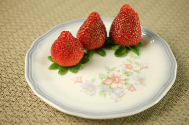 Üç canlı kırmızı çilek zarif bir şekilde dekoratif bir tabakta pastel çiçeklerle süslenmiş taze ve doğal güzellikler sergiliyor. Mutfak ve yaşam tarzı temaları için mükemmel..