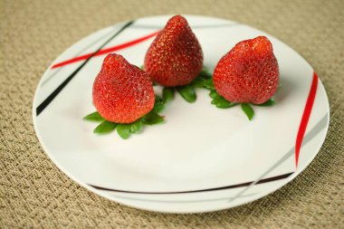 Yeşil yapraklarının üzerinde duran üç dolgun çilek koyu kırmızı ve siyah geometrik aksanlı parlak beyaz bir tabakta güzelce sıralanmış. Taze ve çağdaş görseller için ideal.