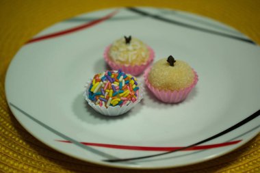 Three brigadeiros on a decorative plate: one with rainbow sprinkles, one coated in coconut flakes, and one sugar-dusted with a clove on top. Perfectly arranged on a vibrant yellow background. clipart