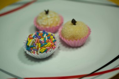 A close-up of three brigadeiros, traditional Brazilian sweets, with colorful sprinkles and a touch of cloves, beautifully arranged in paper cups on a plate. clipart