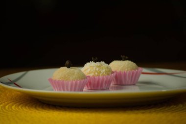 Three coconut truffles topped with cloves, presented in pink paper cups on a patterned white plate, with a warm yellow background enhancing the presentation. clipart