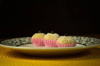 İçinde pembe ambalajlı üç hindistan cevizi şekeri olan bir tabak, her biri özenle bir karanfille kaplanmış. Kompozisyon, canlı sarı bir masa ile tamamlanmış şık siyah-beyaz desenli bir tabağa dayanıyor.