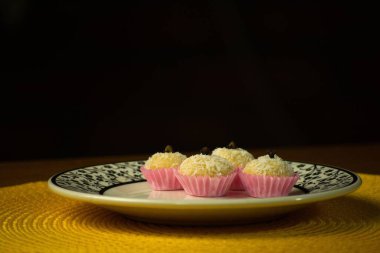 Pembe ambalajlara yerleştirilmiş dört hindistan cevizi şekeri zarif bir tabağa yerleştirilmiş. Her tatlı bir karanfil ile taçlandırılır, sıcak sarı arkaplana karşı büyüleyici bir sunum yaratır..