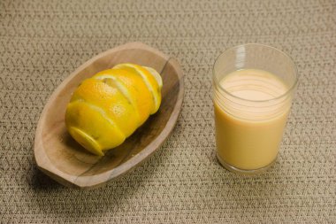 A wooden tray holding a spirally peeled orange, accompanied by a refreshing glass of orange juice, set against a textured beige background for a rustic presentation. clipart