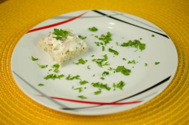 Yumurta salatası ve maydanozla kaplı tek bir parça tam buğday ekmeği, dağınık maydanozla çevrili, canlı sarı arka planda, modern desenli bir tabakta..