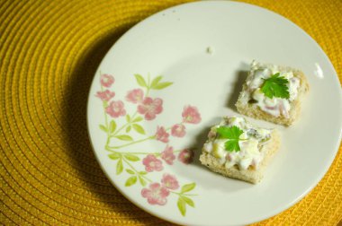 Üzerine krema serpiştirilmiş, maydanozla süslenmiş iki kare mini sandviç. Çiçek desenli bir tabakta sunuluyor..