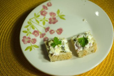 Kremalı salata karışımı ve taze maydanozla kaplı iki kare sandviç. Çiçek tabağında servis ediliyor ve arka planda canlı sarı bir yer örtüsü var..