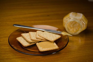 A plate of crackers topped with golden honey, accompanied by a jar of honey and a knife on a wooden table, offering a simple and sweet snack option. clipart