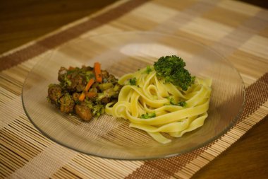 A delicious dish of tagliatelle pasta garnished with broccoli, served alongside sauted chicken and mixed vegetables, presented on a glass plate with a wooden placemat backdrop. clipart